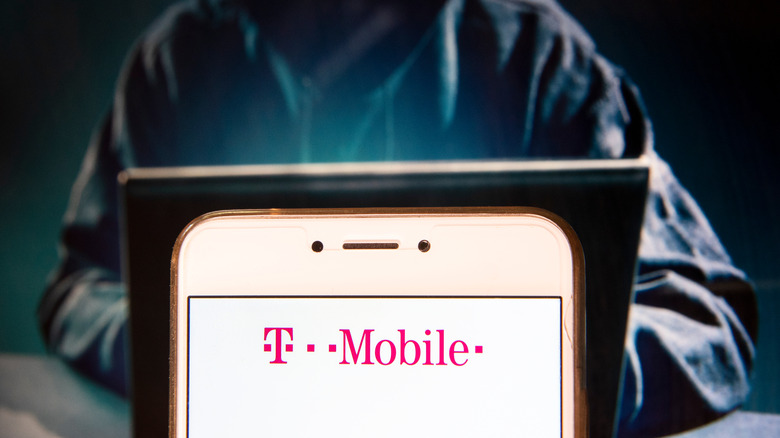 A cybercriminal works at a computer with a phone showing the T-Mobile logo in the foreground