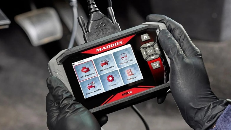 Pair of gloved hands holding a Maddox MR16 OBDII Code Reader that's plugged into a dashboard