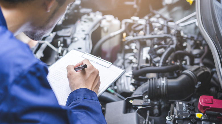 A mechanic holding a checklist and a pen while standing over an engine bay.
