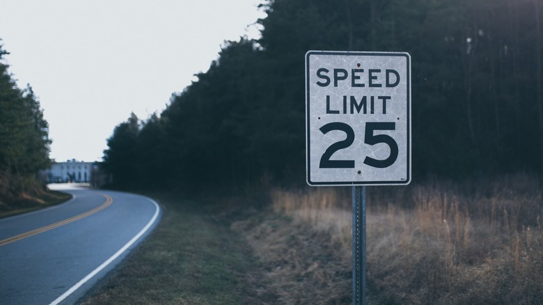 A worn 25-mph speed-limit sign stands beside a quiet, curving rural road lined with trees