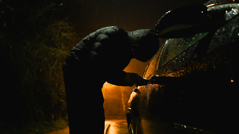A person in all black with a hood attempting to break into a vehicle at night