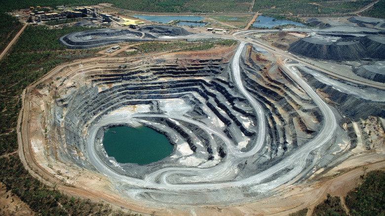 Huge open cast mine set among trees and greenery. Roads lead from the rim, down through terraces, to a lake at the bottom. There is at least one vehicle, but it's almost impossible to see as the scale of the hole is so massive.
