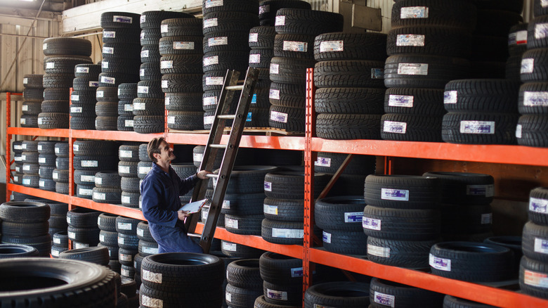 Mechanic holding a new car tire looking upward to shelf of tire inventory