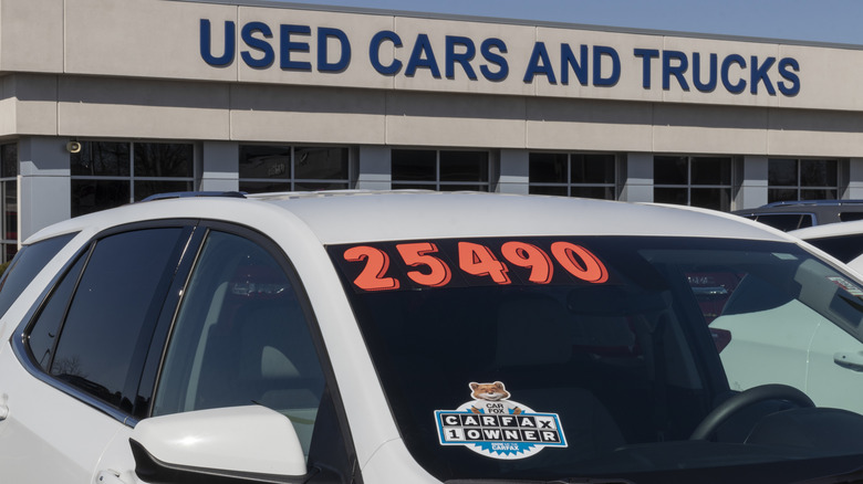 A white car with an orange price tag on its windshield, parked at a used car dealership.