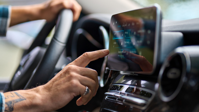 Close up of a car driver with one hand on the steering wheel and the other hand operating the car's dashboard screen.