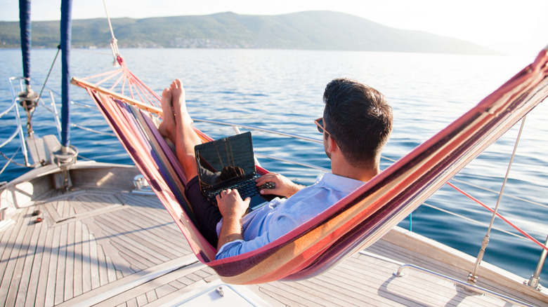 A man on a laptop sitting in a hammock that's on a sail boat with land in the background.