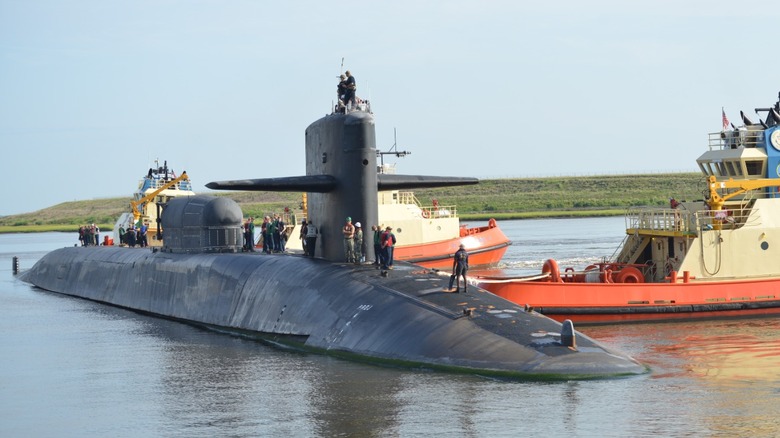 U.S. submarine with two tug boats in an inlet while Navy personnel walk along the sub's hull.