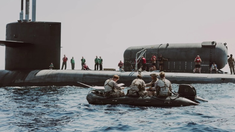 U.S. Marines sit in rigid hull boat with crew standing on submarine hull watching in the background.
