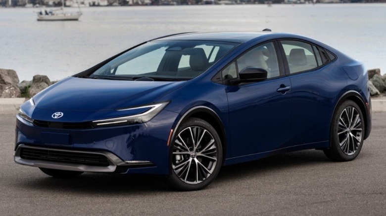 A Reservoir Blue Toyota Prius Limited parked on a paved surface with a body of water in the background.