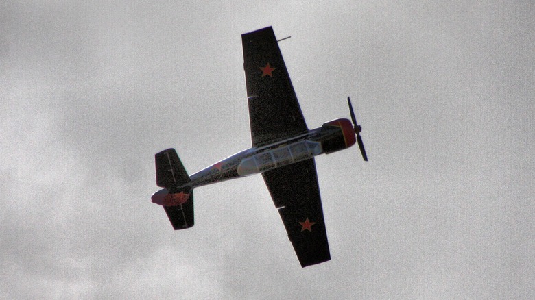 Black Yak-52 flying upside-down over cloudy skies showing red stars on wings