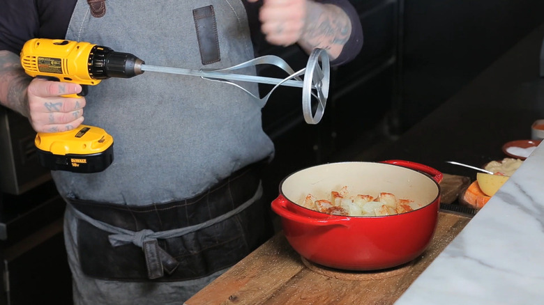 A person holds a DeWalt drill with a large mixing attachment over a pot of food on a kitchen counter.