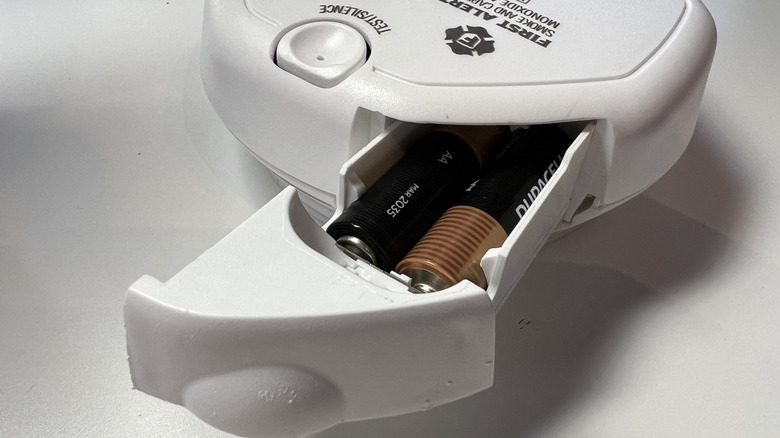 A smoke alarm with its battery compartment open revealing two AA disposable batteries