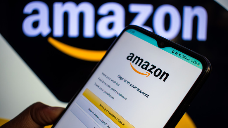 A man holds a smart phone with the Amazon app sign-in screen displayed, with the Amazon logo displayed behind the phone