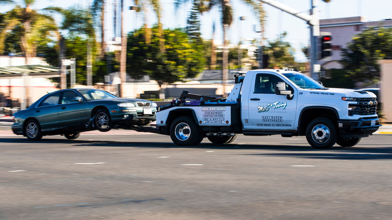 A white tow truck towing a green sedan with its front wheels off the ground through a junction.