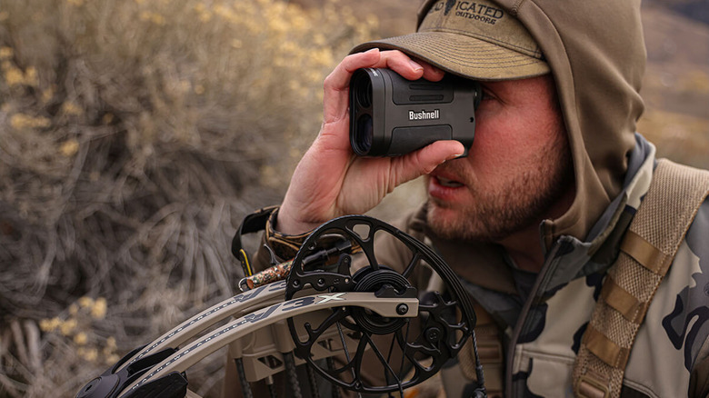 A man in camouflage using a rangefinder with his bow underneath him.