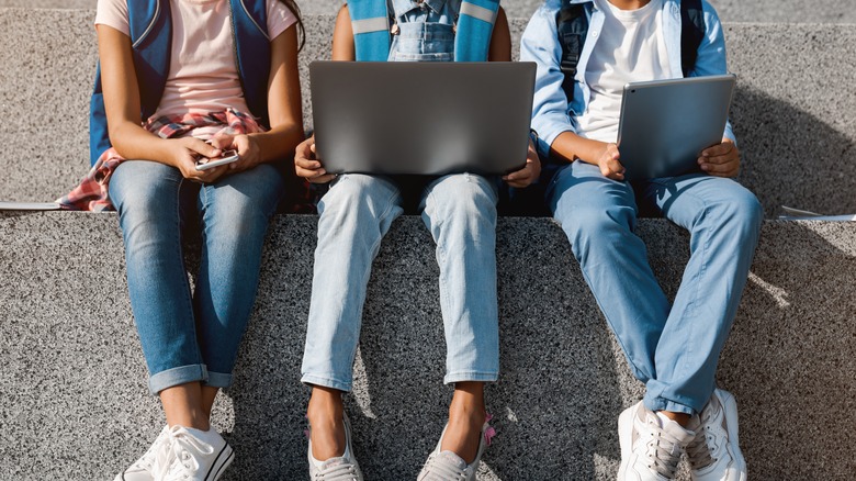 Cropped image of school kids preteen boys and girls elementary middle pupils sitting on the stairs and using digital devices wireless gadgets laptop tablet smartphone outdoors at the street.