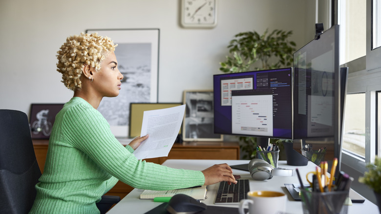 Side view of a woman in a green top holding a document while using a desktop computer.