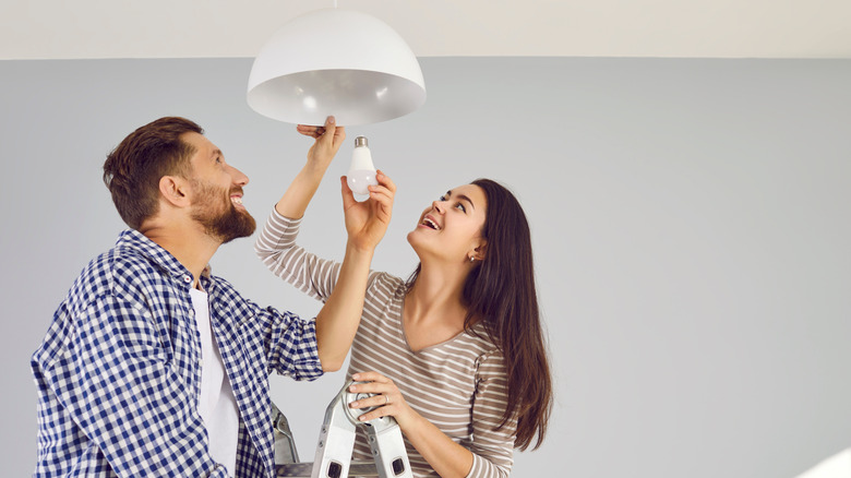 A man and a woman standing on a step ladder at home placing an LED lightbulb in a white lamp on the ceiling