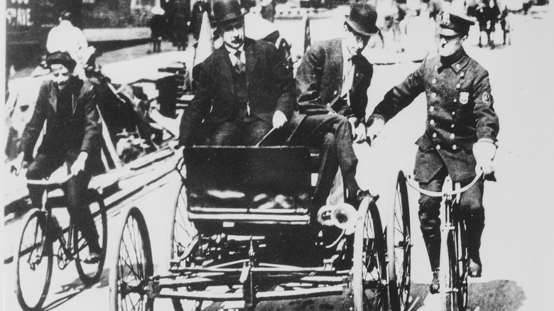 Traffic violator driving a 1900-vintage car being stopped by a policeman on a bicycle.