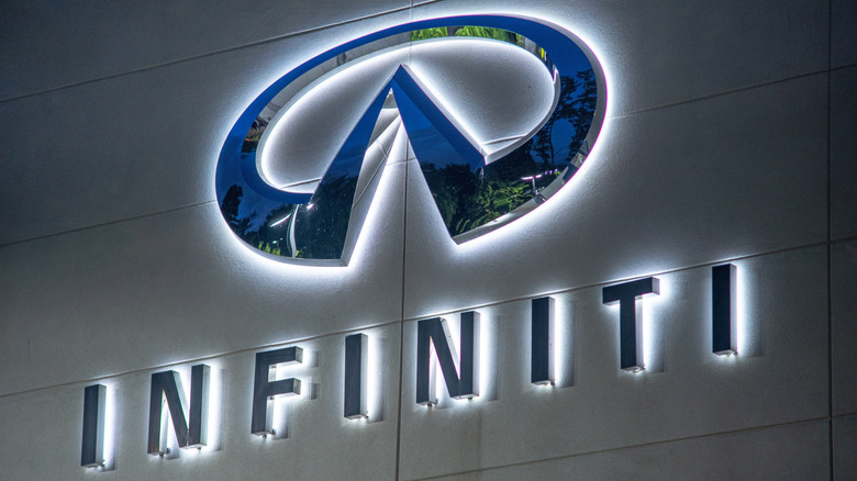 An illuminated bronze Infiniti logo mounted to the side of a building after sunset.