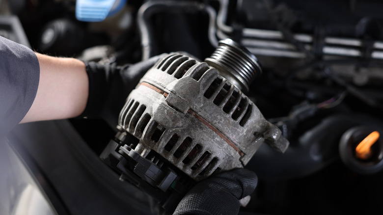 A mechanic holding a car generator above a car's engine bay.