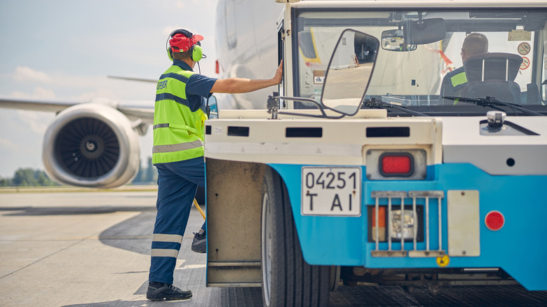 Two ground crew workers in hi-vis, one inside and one outside a tow vehicle, facing a passenger plane in front of them