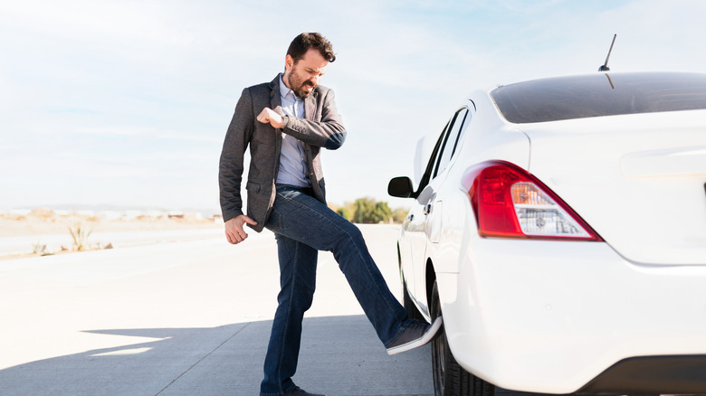 A man in white shirt, gray blazer and denim pants kicking one of his white sedan's rear tires on the side of the road. He looks angry.