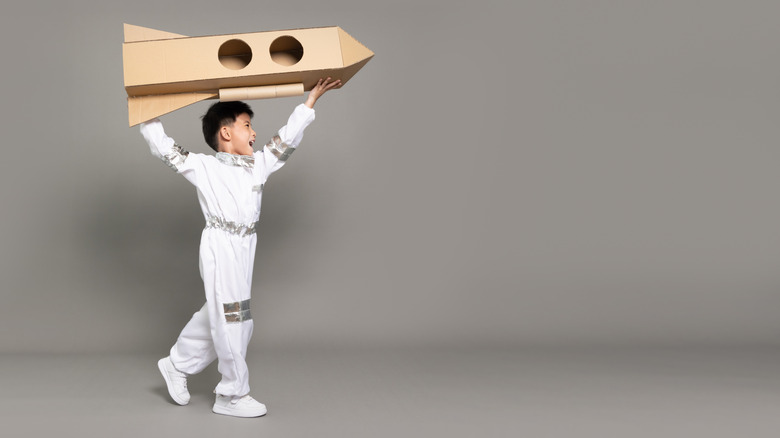 Child playing with a cardboard rocket on grey background.