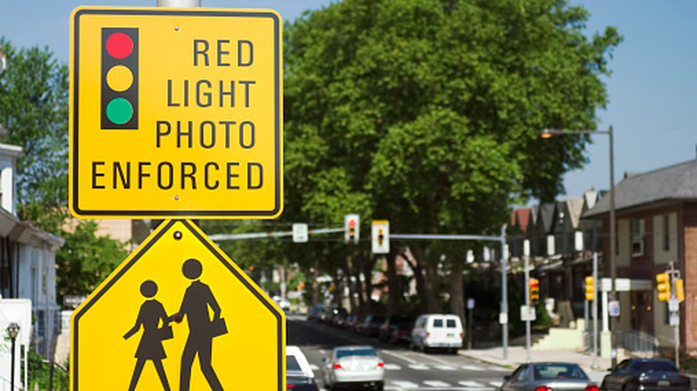 A "Red Light Photo Enforced" sign stands in front of a traffic light warning drivers. Cameras at the intersection take pictures of cars that run through a red light.