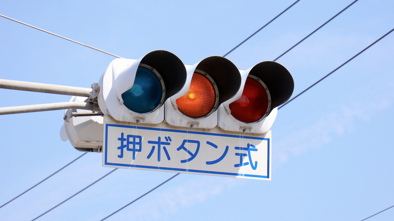 A traffic light signal that's blue, amber, and red. It's on a light blue sky background with a sign underneath it saying push button type.