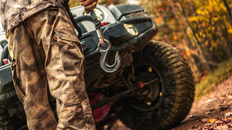 A person wearing camo clothing stands in front of an off-road SUV, holding a rope and a hook.