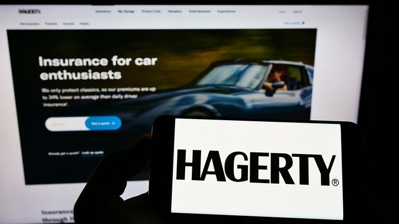 A smartphone displaying a Hagerty logo in front of a computer monitor.