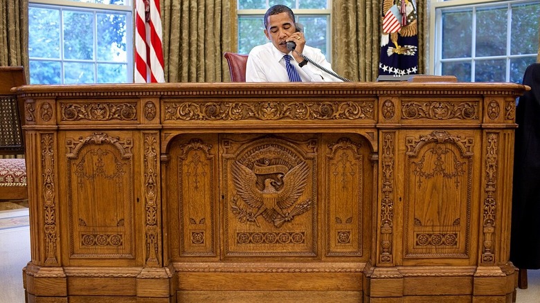 Barak Obama on the phone sitting at the Resolute desk