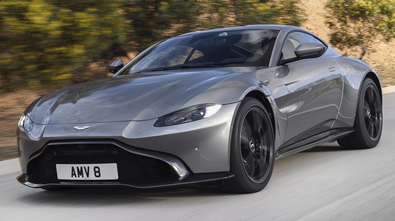 A silver Aston Martin Vantage coupe moving on a road with a scenic mountain backdrop.