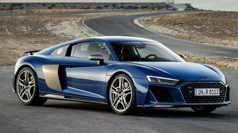 A blue Audi R8 sitting confidently on a racetrack, backed by a winding road and mountain views.