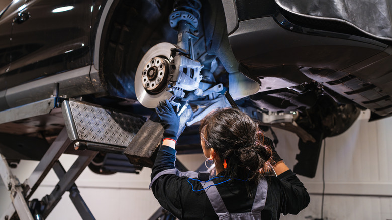 A female mechanic inspecting a car's brake assembly with a flashlight in a shop.